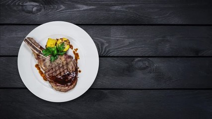 Rib-eye steak with corn rotating on a white plate. Top view on black wooden background. Copy space for your text.
