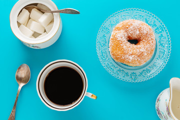 A cup of coffee, a plate with sugar coated doughnut and other breakfast objects, on blue background.