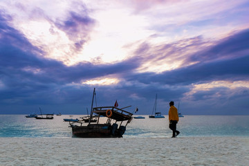 Silhouette of a man goes in the direction of a wooden boat on the beach against the backdrop of a beautiful sunset