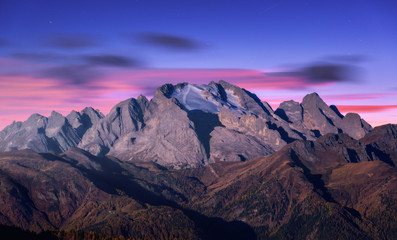Mountain peak lighted by moonlight in autumn at night in Dolomites, Italy. Beautiful landscape with mountains, forest on hills, purple sky with pink clouds, stars at dusk. Italian alps. High rocks  - obrazy, fototapety, plakaty