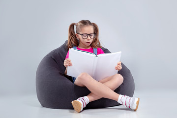 Young girl in glasses reading a book on a gray bean bag