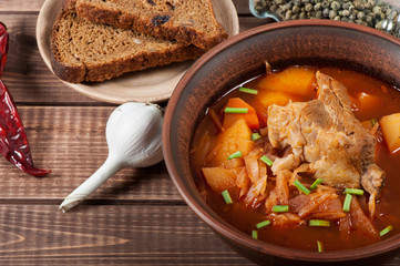 Traditional Ukrainian borsch with meat on the edge, in a brown clay bowl, on a wooden background.