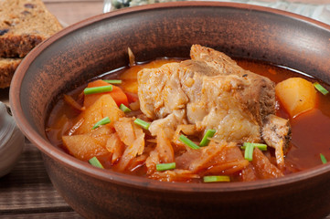 Traditional Ukrainian borsch with meat on the edge, in a brown clay bowl, on a wooden background.