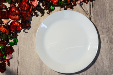 Empty white plate on a wooden background decorated with dry flower petals. View from above.