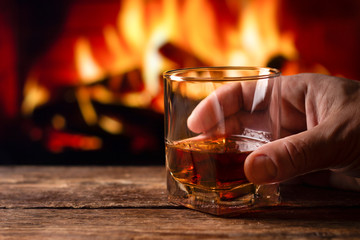 A glass of whiskey in a man hand. Blur burning fireplace background.