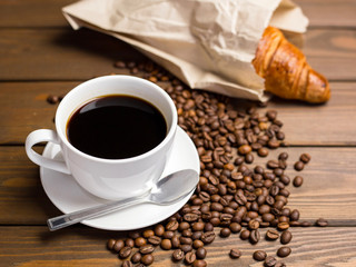 Coffee cup, beans and croissant in paper packaging on wooden background on the table. Perfect breakfast in the morning. Rustic style