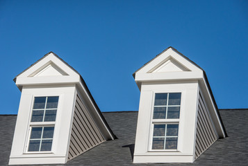 Double white dormer sash window with blue sky background on a gable roof with vinyl siding on a...