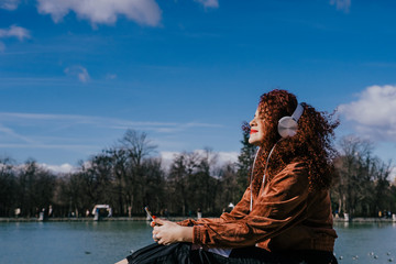 Young woman with afro hair listening to music with headphones at the edge of a small lake. Full of positive energy, fun and carefree. Lifesytle.