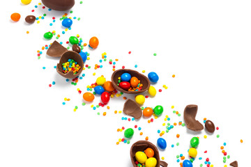 Fototapeta premium Broken and whole chocolate Easter eggs, multicolored sweets on white background. Concept of celebrating Easter, Easter decorations, search for sweets for Easter Bunny. Flat lay, top view. Copy Space