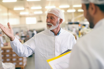 Two Caucasian workers in protective suit talking and smiling while standing in food factory.