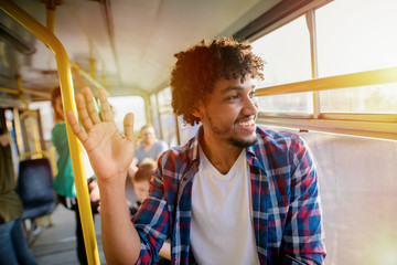 Young modern Afro-American man standing in a bus while looking through the window and waving to his...