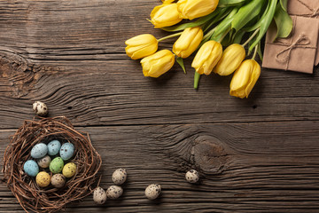 Yellow tulips in a paper bag, a nest with Easter eggs on a wooden background. Top view with copy space.