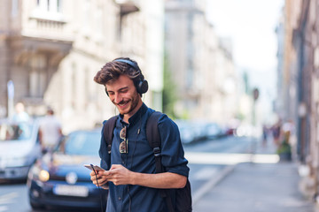 Positive young man holding phone and listening music on headphones at street