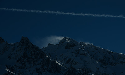 Intentionally underexposed dark image of wind blowing over snow capped mountain peaks. Italian Alps, Passo Tonale, Val Camonica, Italy. Plane trail. 