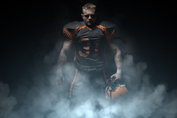 American football player on a dark background in smoke in black and orange equipment. © bondarchik