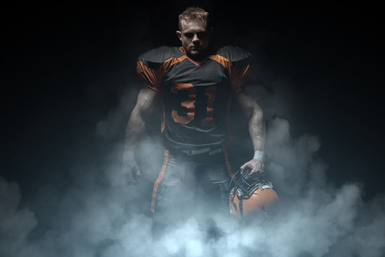 American football player on a dark background in smoke in black and orange equipment.
