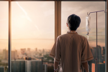 Asian man hand of a patient standing with medical drip or IV drip in hospital ward and look out at window with sunrise time, Selective focus, Healthcare concept.