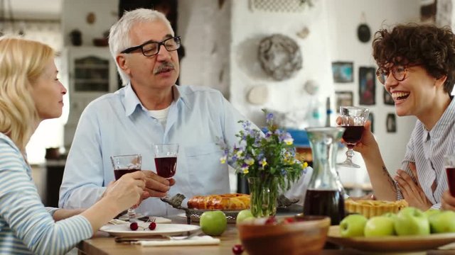 Pan shot of members of family sitting at dinner table holding glasses with red wine and talking