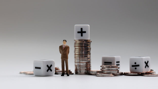 A miniature businessman standing with a pile of coins and an arithmetic symbol cube.
