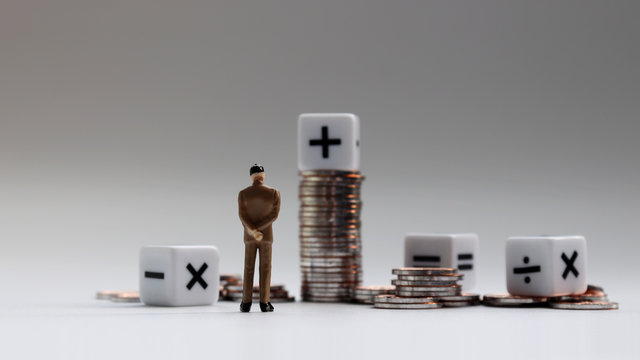The back of a miniature businessman standing with a pile of coins and an arithmetic symbol cube.