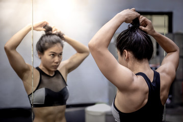 Asian athlete girl in sportswear looking into the mirror while tying her hair in fitness gym....