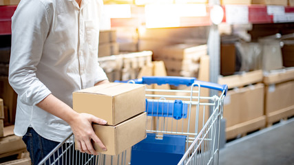 Male shopper putting cardboard box into trolley cart near product shelves in warehouse. Shopping in department store or Inventory delivery concepts