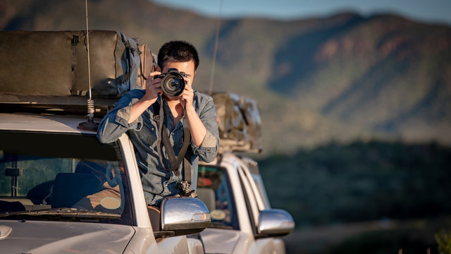 Young Asian man traveler and photographer sitting on the car window taking photo on road trip in Namibia, Africa. Travel photography concept