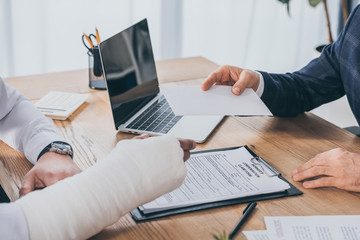 cropped view of businessman sitting at table and giving paper sheet to worker with broken arm in office, compensation concept - obrazy, fototapety, plakaty