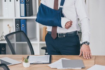cropped view of worker with broken arm in bandage standing near table in office, compensation...