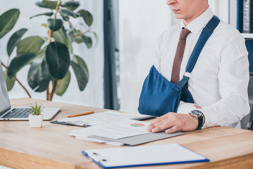 cropped view of worker with broken arm in bandage sitting at table and reading documents in office,...