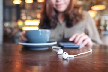 Closeup image of a woman drinking coffee while using mobile phone to listen music with earphone on wooden table in cafe