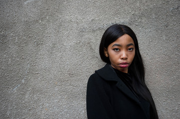 20s worried long hair young African woman portrait in black dress with grey cement wall