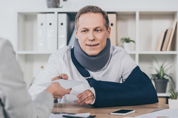 woman giving worker in neck brace and arm bandage envelope over table in office, compensation concept