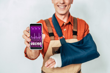 cropped view of repairman with broken arm and bandage showing smartphone at white background