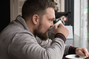 Handsome bearded man in light pullover drinking espresso, looking at window of the coffee shop. Close up os serious man freelancer is sitting at the table of the coffee shop
