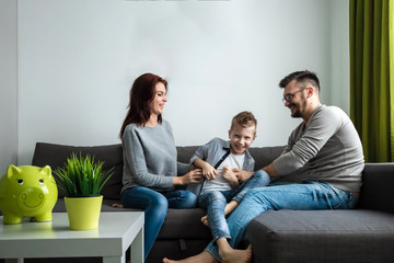 Happy parents and son having fun, tickling sitting together on the sofa, cheerful couple laughing,...
