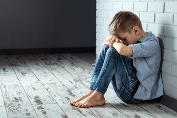 A young boy sits alone with a sad feeling at school near the wall. Offended child abandoned in the corridor and bent against a brick wall. Bullying, discrimination with copy space.