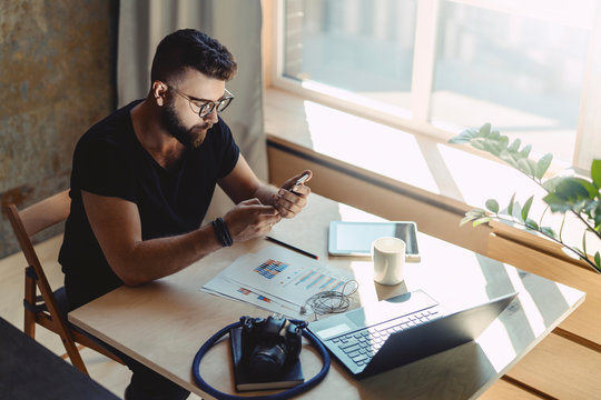 Man Millenial In Glasses Sits At Table In Front Of Laptop And Uses Smartphone, Reads Message From Business Partner.