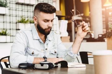 Portrait of young bearded male writer. Guy sits with notebook in cafe at table, holds pencil in his hand and reads book. Journalist writes article for newspaper. Student doing homework, reads synopsis