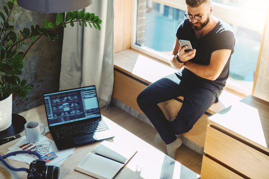 Attractive hipster guy sits home on windowsill, reads text messages, sends e-mail, browses social networks, uses his smartphone and fast internet connection. On table is laptop, camera, paper notebook