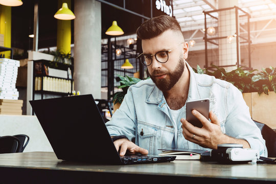 Hipster man sits in cafe, uses smartphone, works on laptop. Businessman reads an information message in computer. Freelancer works outside office, teleworking. Online marketing, education for adult.