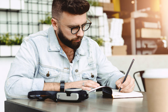 Young bearded hipster man sits in cafe at table, writes in notebook. Journalist writes information in notepad. On table cup of coffee, camera. Man is working, studying, learning. Education for adult.