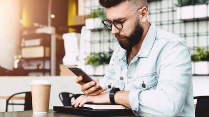 Serious hipster man reading message on smartphone while sitting in cafe at table. Guy checks mail through smartphone while relaxing in restaurant. Businessman checks information using phone. Education