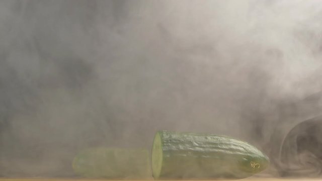 A man cuts a farmer's juicy cucumber with a knife in haze and evaporation, black background, close-up, vegetable, slow motion
