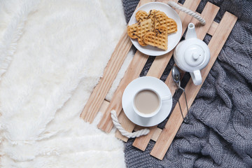 Cozy breakfast in bed, cup of coffee and heart shaped waffles on wooden tray on white and gray cozy blanket, the concept of home comfort, copy space