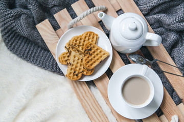 Fototapeta na wymiar Cozy breakfast in bed, cup of coffee and heart shaped waffles on wooden tray on white and gray cozy blanket, the concept of home comfort, copy space