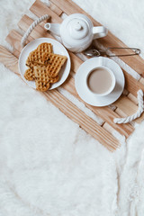 Obraz na płótnie Canvas Cozy breakfast in bed, cup of coffee and heart shaped waffles on wooden tray on white and gray cozy blanket, the concept of home comfort, copy space