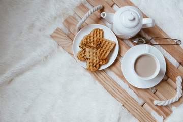 Cozy breakfast in bed, cup of coffee and heart shaped waffles on wooden tray on white and gray cozy blanket, the concept of home comfort, copy space