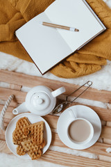 Cozy breakfast in bed, cup of coffee and heart shaped waffles on wooden tray on white cozy blanket with yellow sweater and notebook, the concept of home comfort, copy space