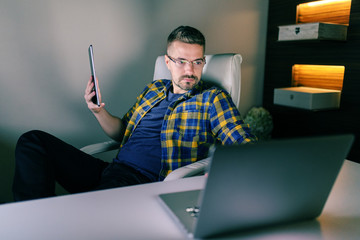 Young unshaven employee holding one hand on laptop and in other holding tablet while sitting in the...